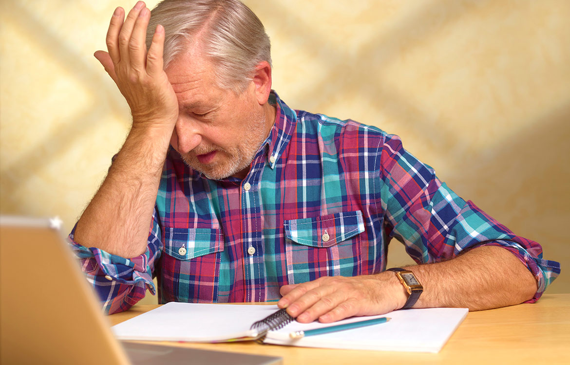 older man sitting at desk in pain with his hand on forehead