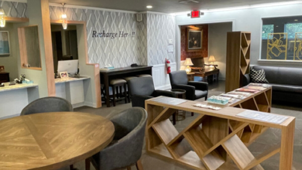 Reception area with a wood check-in desk, a round table with cushioned chairs, a lounge seating group, and a "Recharge Here!" sign on a patterned accent wall.