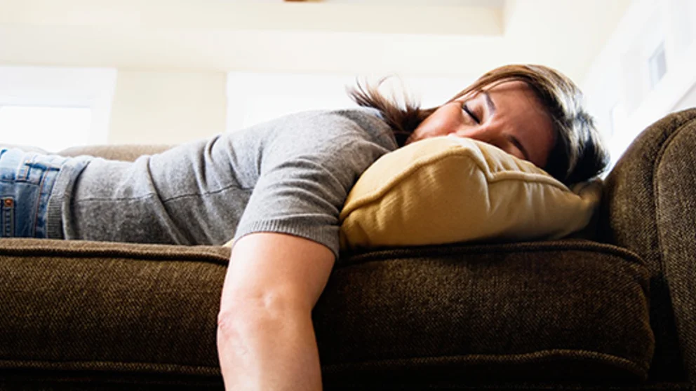 A middle-aged woman lies on a couch exhausted. Fatigue is caused by many things including poor sleep hygiene habits, lack of exercise and some conditions.