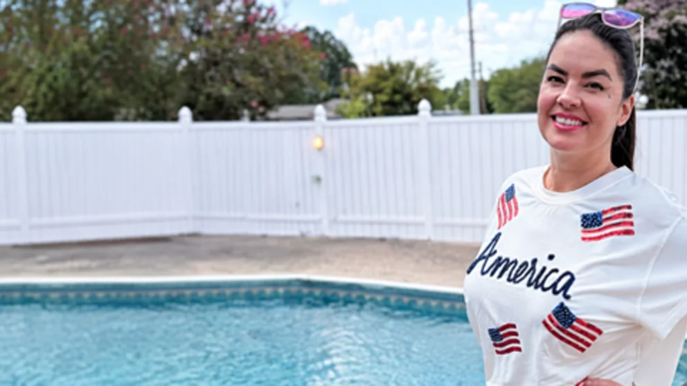 Smiling woman with colorful tattoos standing beside a backyard swimming pool.