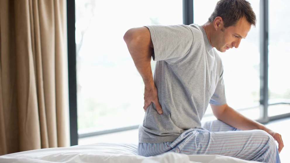 Middle aged man sitting on the edge of his bed, gripping his lower back.